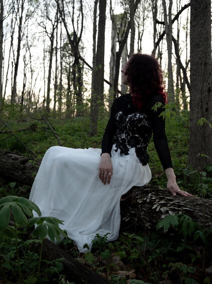 Photo of Emi sitting on a log in a forest. Her red-dyed hair is obscuring her face as she turns to look toward the sunset. She is wearing a white gown with black lace.